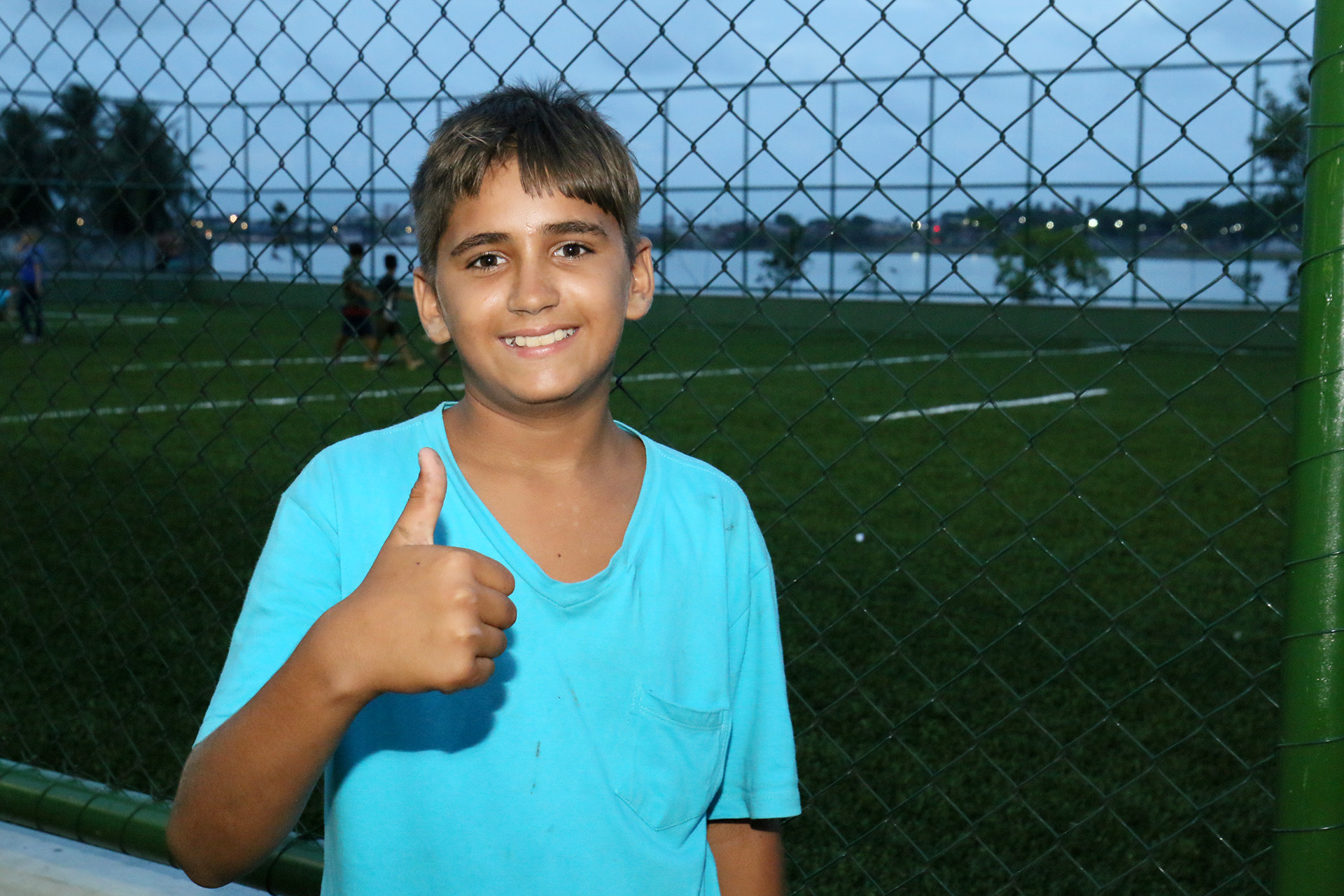 garoto sorrindo para a foto e fazendo sinal de positivo com campo de futebol ao fundo